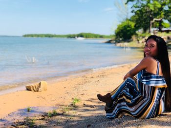 Portrait of young woman sitting on shore at beach against clear sky