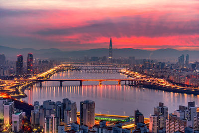 Illuminated buildings by river against sky during sunset