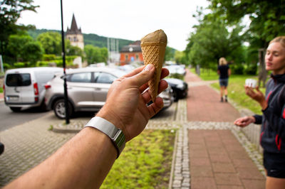 Cropped hand of man holding ice cream cone against woman standing on footpath in background