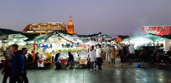 People walking on illuminated street market in city against sky