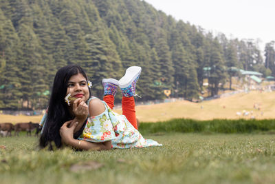 A cheerful smiling indian girl lying on green grass field looks away with a flower in hand