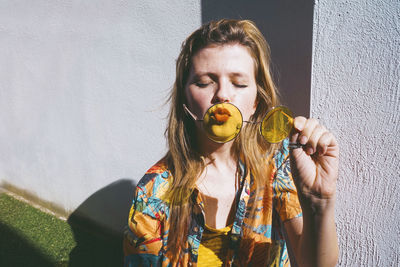 Woman with eyes closed holding yellow sunglasses by wall on sunny day