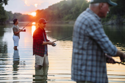 Side view of male friends fishing while standing in lake during sunset