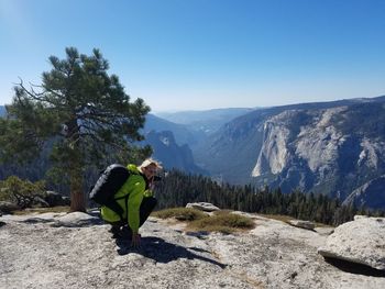 Mature woman photographing while crouching on mountain against clear sky