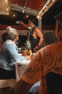 Ethnic male taking selfie photo on cellphone with group of friends gathering at table of terrace for party