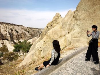 Rear view of couple sitting on rock formation against sky