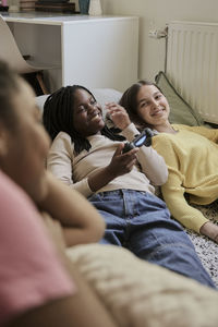 Happy teenage girl playing video game while lying down with female friend in bedroom