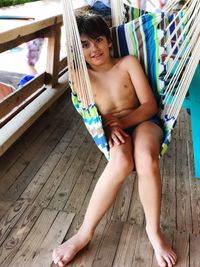 High angle portrait of smiling shirtless boy sitting in hammock at porch