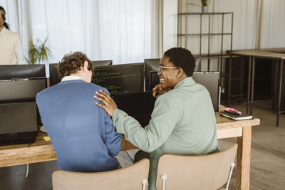 Rear view of smiling female programmer sitting with male colleague at computer desk in office