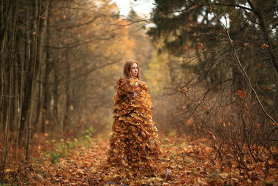 Portrait of young woman covered with leaves standing at forest during autumn