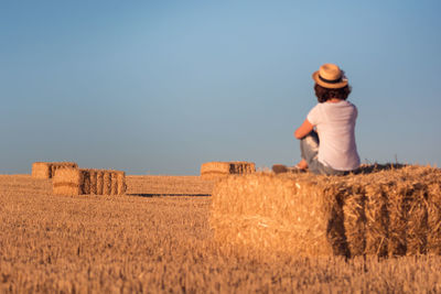 Woman sitting on hay bale against clear sky
