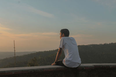Man sitting on retaining wall against sky during sunset