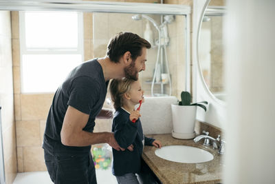 Side view of father watching daughter brushing teeth at sink in bathroom