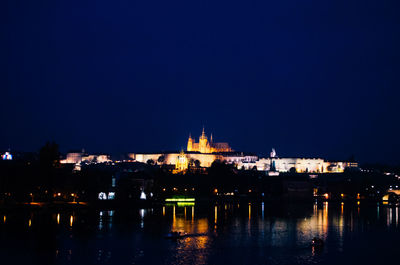 Illuminated buildings by river against blue sky at night