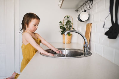 European child of 4 years old washes glass in sink on his own, bright kitchen in real interior, 