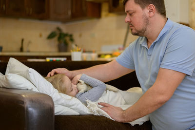 Dad touches the forehead of his sick child, who is lying on the couch, covered with a white blanket