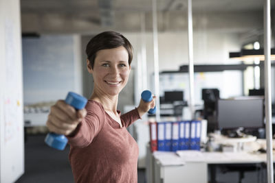 Smiling businesswoman in office exercising with dumbbells