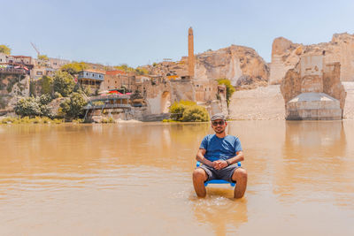 Portrait of man sitting on chair in lake against houses