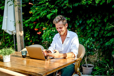 Middle aged businessman in smart casual clothes smiling and scrolling data on cellphone while sitting at table with laptop during work in street restaurant