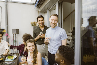 Happy male friends with group of friends enjoying drinks during dinner party in balcony