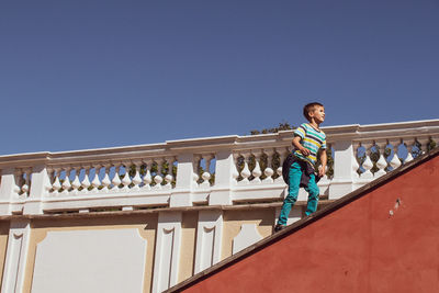 Low angle view of boy standing by railing against clear blue sky