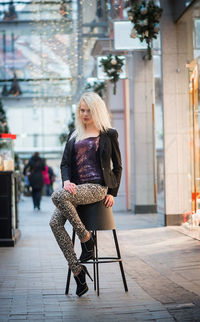 Full length portrait of woman sitting on stool against building in city