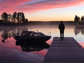 Rear view of silhouette person standing at pier over lake against cloudy sky during sunrise