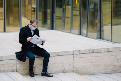 Full length of businessman wearing mask reading newspaper while sitting outdoors