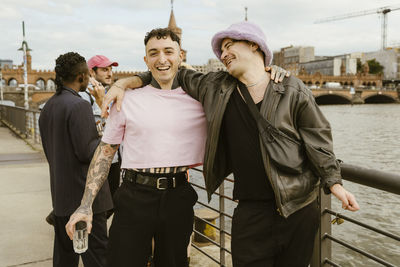 Portrait of happy non-binary person standing with arm around gay friend near railing on promenade in city