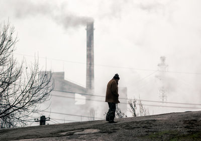 Rear view of man standing by factory against sky