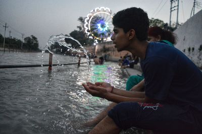 Side view of teenage boy splashing water while sitting at river