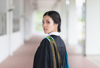 Portrait of young woman wearing graduation gown standing in corridor at university