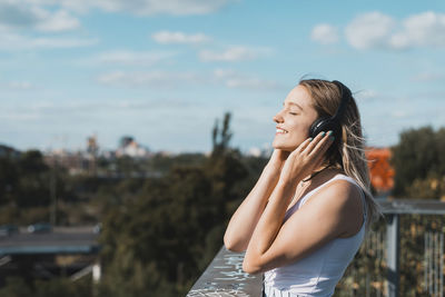 Side view of smiling young woman listening music by railing against sky
