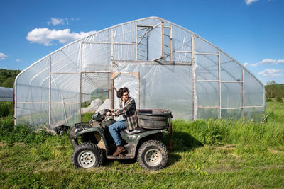 Young farmer on off road vehicle in green sunny field by vegetable greenhouse