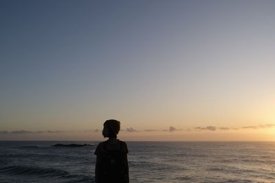 Rear view of woman standing at beach against clear sky during sunset