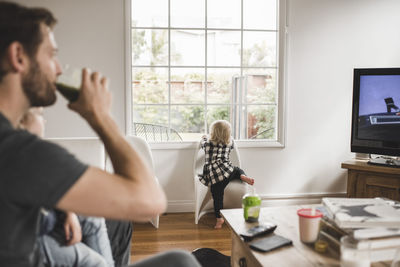 Girl climbing chair against window with father watching tv in foreground at home