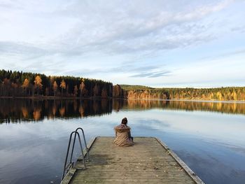 Rear view of woman sitting on pier over lake against sky