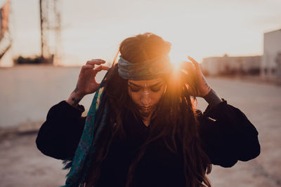 Attractive woman with ethnic rite makeup and tattoo with closed eyes while standing on street and touching head against blurred urban environment during sunset