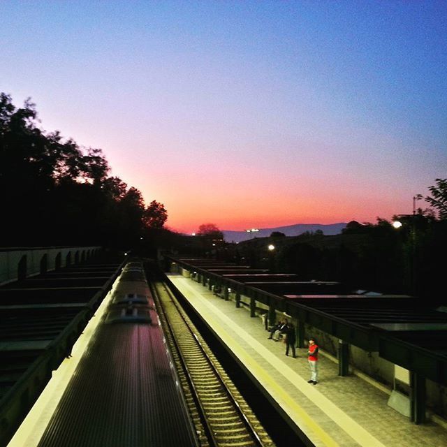 railroad track, transportation, rail transportation, clear sky, sunset, illuminated, the way forward, public transportation, tree, railroad station, railroad station platform, dusk, sky, copy space, high angle view, diminishing perspective, travel, vanishing point, blue, built structure