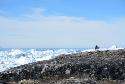 Rear view of man sitting on rock formation by glacier against sky