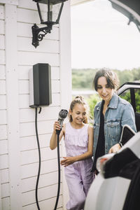 Smiling daughter with mother charging electric car outside house