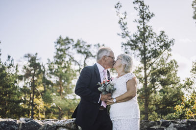 Affectionate newlywed senior couple kissing on mouth against forest on wedding day