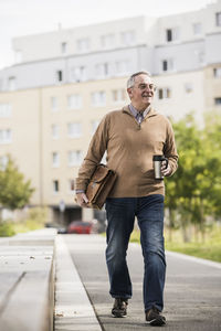 Happy senior man holding bag and insulated drink container on footpath