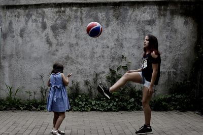 Sisters playing with ball while standing on footpath against wall