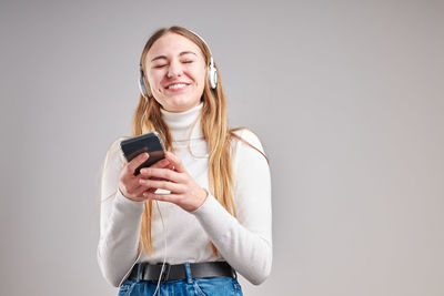 Smiling teenage girl holding mobile phone listening music against gray background