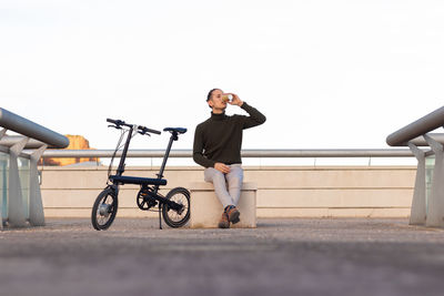 Young man sitting on bench with battery bike on the side taking a break driking a cup of coffee