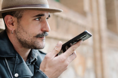 Side view of bearded male with mustache in hat and leather jacket using mobile phone and sending voice message on blurred background of building looking away