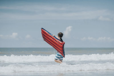 Teenage boy with pool raft walking in sea against sky during sunny day
