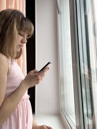 A woman in a pink dress standing at the window looks at the screen of the phone.