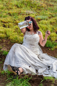 Young woman pouring liquid on wedding dress while sitting on field